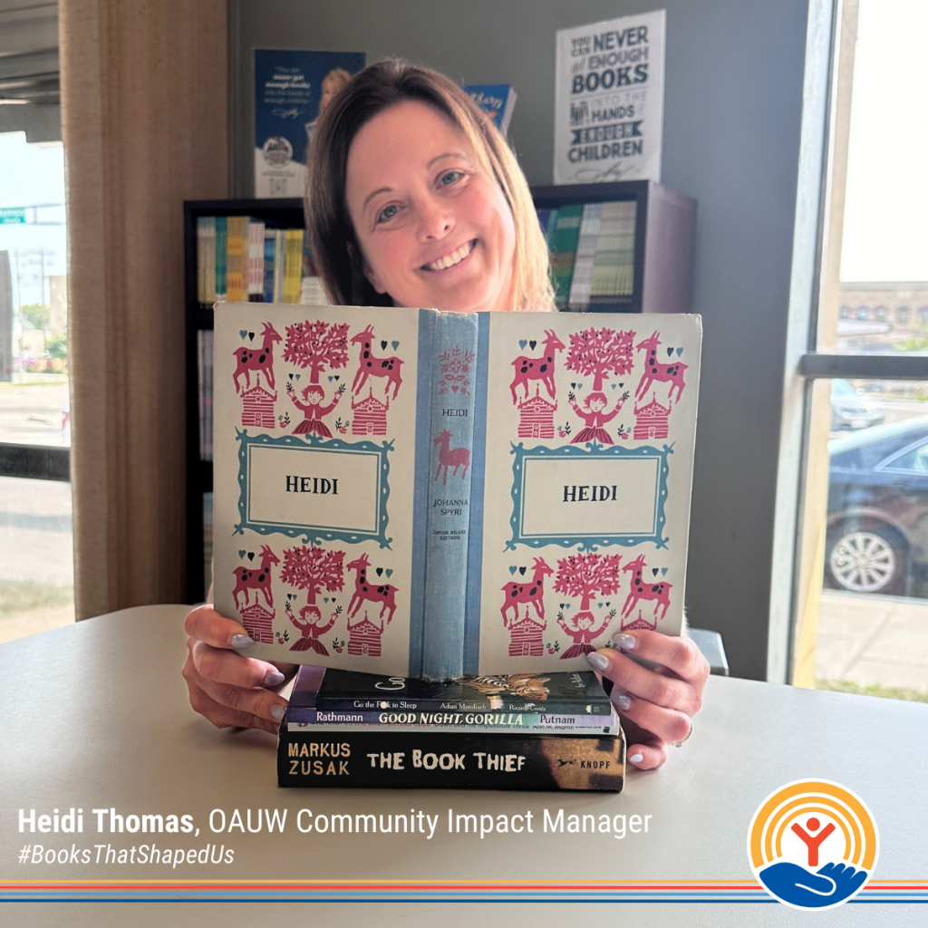 Heidi Thomas, OAUW Community Impact Manager, holds a copy of the book Heidi by Johanna Spyri. The book is open, and a stack of other books including Good Night, Gorilla and The Book Thief is visible on the table in front of her. She is smiling in a cozy, well-lit room filled with books.