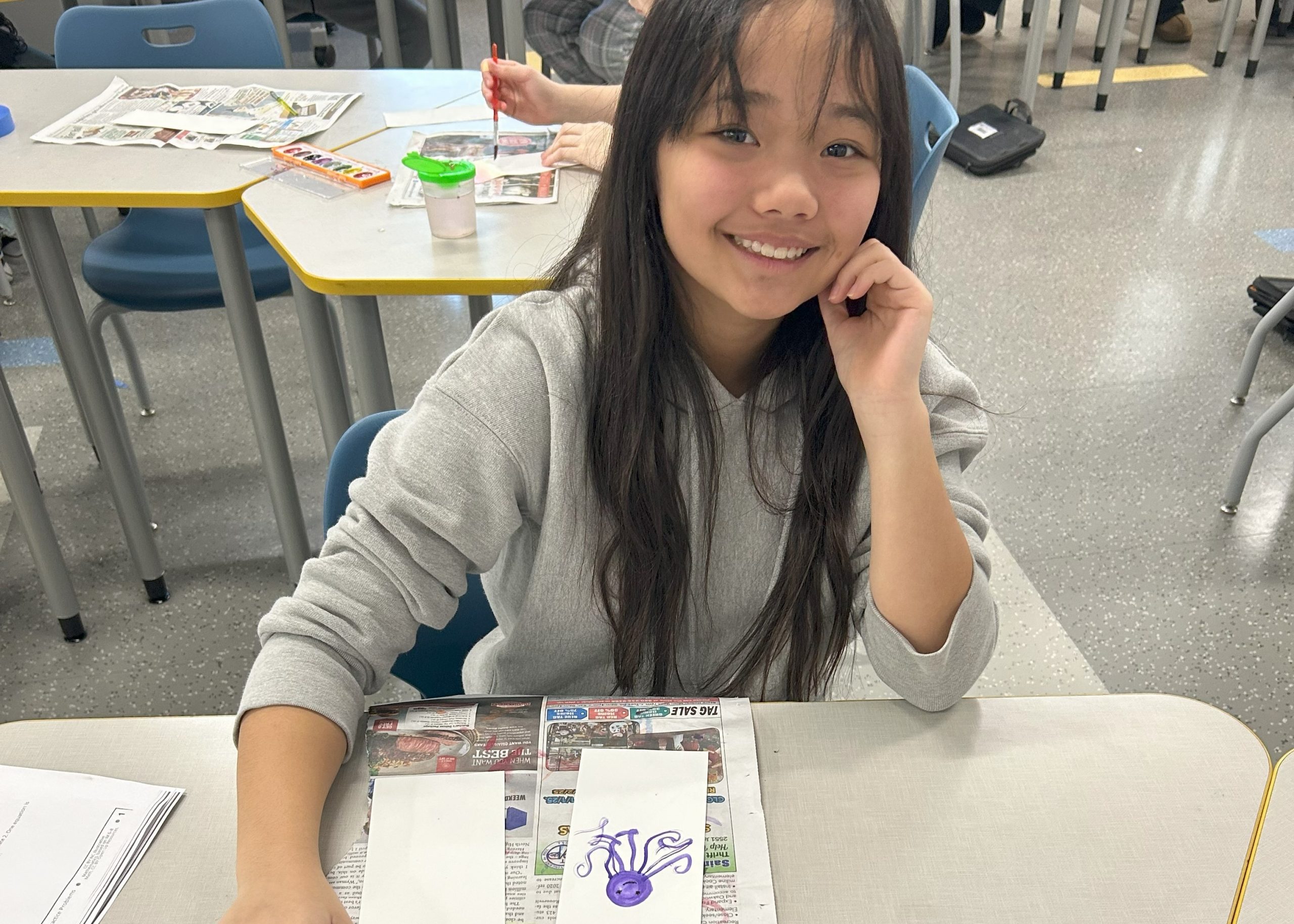 A smiling middle school student with long dark hair shows her watercolor painting of a purple octopus, created during a classroom art project.